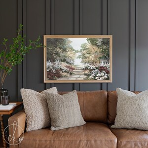 Puede incluir: Una pintura enmarcada de un camino de jard&iacute;n con &aacute;rboles y flores. La pintura est&aacute; colgada en una pared gris oscuro sobre un sof&aacute; de cuero marr&oacute;n con dos almohadas.