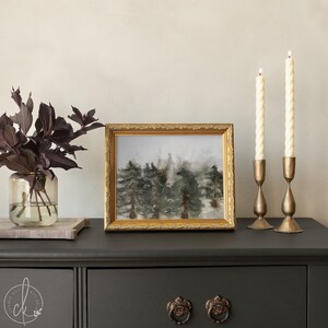 May include: A framed landscape painting with a gold ornate frame, depicting a forest scene in muted tones. Two brass candle holders with white candles are on the right. A glass vase with dark purple leaves is on the left.