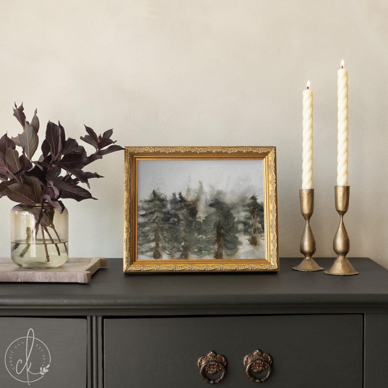 May include: A framed landscape painting with a gold ornate frame, depicting a forest scene in muted tones. Two brass candle holders with white candles are on the right. A glass vase with dark purple leaves is on the left.