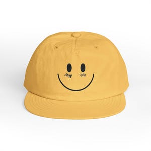 Può includere: Cappellino da baseball giallo senape con un design a faccina sorridente. Il cappellino ha occhi neri, una bocca curva e la scritta "Always Tired" sopra gli occhi. Presenta una visiera curva e un bottone sulla parte superiore.