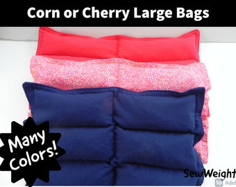 May include: Three large heat packs, one red, one pink with white polka dots, and one navy blue. The heat packs are stacked on top of each other. The text "Corn or Cherry Large Bags" is at the top of the image. The text "Many Colors!" is in a starburst shape at the bottom left of the image. The text "SewWeighty" is at the bottom right of the image.