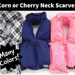 May include: Three neck scarves in a row. The first is black and white plaid, the second is solid navy blue, and the third is pink with a white floral pattern. Text at the top says "Corn or Cherry Neck Scarves". A starburst graphic says "Many Colors!"