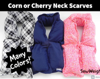 May include: Three neck scarves in a row. The first is black and white plaid, the second is solid navy blue, and the third is pink with a white floral pattern. Text at the top says "Corn or Cherry Neck Scarves". A starburst graphic says "Many Colors!"