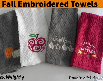 May include: Embroidered fall-themed kitchen towels in gray, white, and red. Designs include a pumpkin, apple, the word "hello", and "Share the Harvest". The towels have a textured waffle weave.