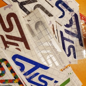 May include: A stack of various colored vinyl stickers with the word "STS" printed on them. The stickers are on a white gridded backing paper. The text "StrongGrip Transfer" is visible on one of the stickers.