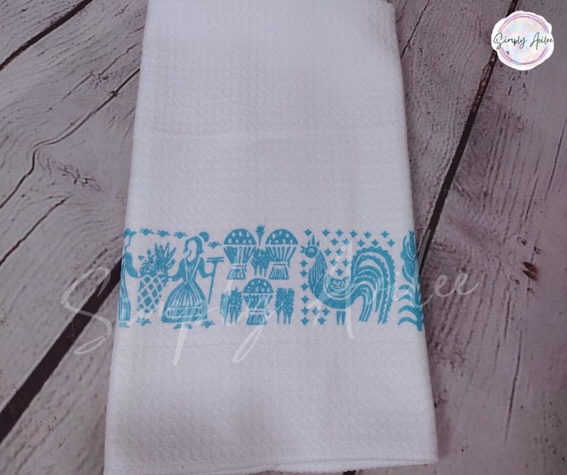 May include: White kitchen towel with a textured surface and a turquoise border featuring a rooster, woman, and pineapple design. The towel is folded and displayed against a wooden background.