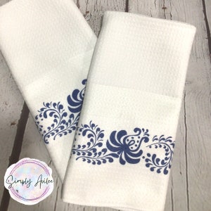May include: Two white kitchen towels with a blue floral design along the bottom edge. The towels are folded and stacked on top of each other.