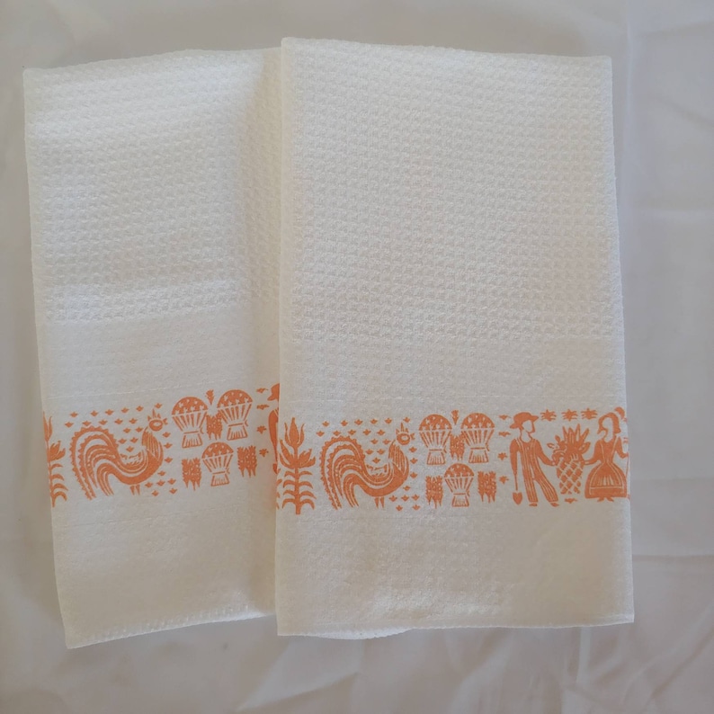 May include: Two white waffle-weave kitchen towels with an orange border featuring rooster and folk art designs. The towels are folded and displayed on a white surface. The towels are made of a soft, absorbent material.