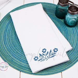 May include: A white kitchen towel with a blue floral design. The towel is folded and placed on a blue and green circular placemat.