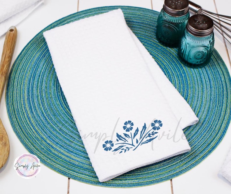 May include: A white kitchen towel with a blue floral design. The towel is folded and placed on a blue and green circular placemat.