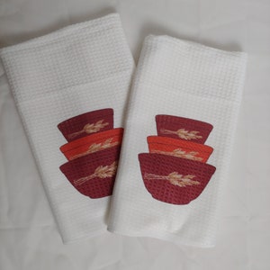May include: Two white waffle-weave kitchen towels. Each towel features a design of stacked bowls in shades of red and orange, with wheat stalks. The towels are clean and neatly folded, suitable for kitchen use.