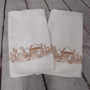 Puede incluir: Dos toallas de cocina blancas con un diseño de champiñones y hojas marrones. Las toallas están dobladas y colocadas sobre una superficie de madera.