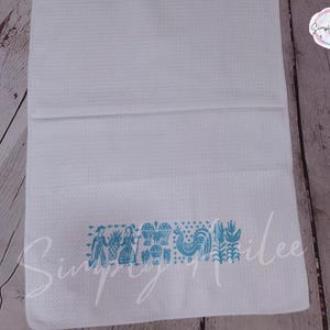 May include: White waffle-weave kitchen towel with a turquoise folk art design. The design includes figures, a rooster, and floral elements. The towel is folded and displayed on a wooden surface.