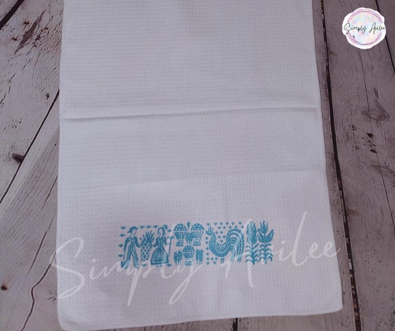 May include: White waffle-weave kitchen towel with a turquoise folk art design. The design includes figures, a rooster, and floral elements. The towel is folded and displayed on a wooden surface.