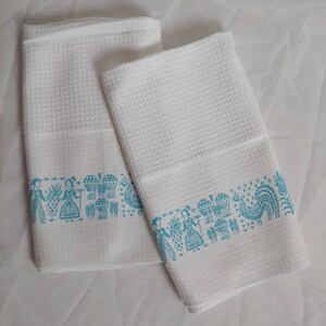 May include: Two white cotton towels with a textured weave. Each towel features a turquoise border with a repeating pattern of figures, baskets, and a rooster. The towels are folded and displayed on a white surface.