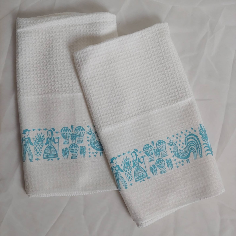 May include: Two white cotton towels with a textured weave. Each towel features a turquoise border with a repeating pattern of figures, baskets, and a rooster. The towels are folded and displayed on a white surface.