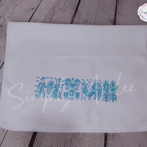 May include: White waffle-weave kitchen towel with a turquoise folk art design. The design includes figures, hot air balloons, a rooster, and floral elements. The towel has the text "Simply Ailee" in a script font.