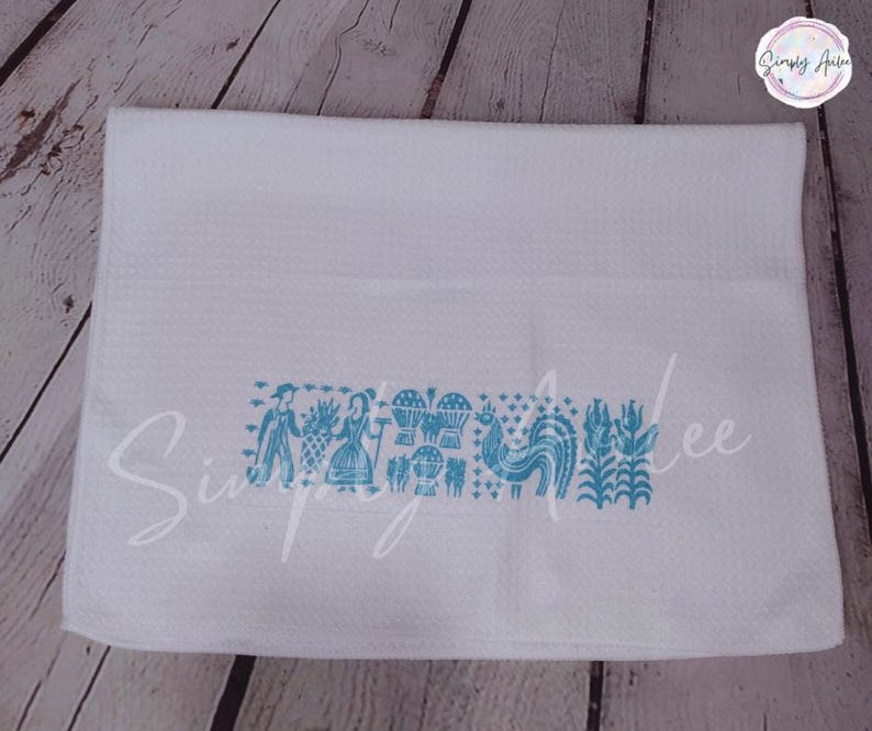 May include: White waffle-weave kitchen towel with a turquoise folk art design. The design includes figures, hot air balloons, a rooster, and floral elements. The towel has the text "Simply Ailee" in a script font.