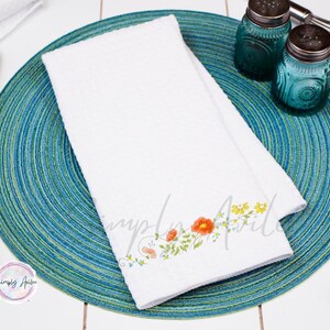 K&ouml;nnte beinhalten: Ein wei&szlig;es K&uuml;chentuch mit einem Blumendesign mit orangefarbenen, gelben und gr&uuml;nen Blumen. Das Tuch liegt auf einem blauen und gr&uuml;nen runden Untersetzer.