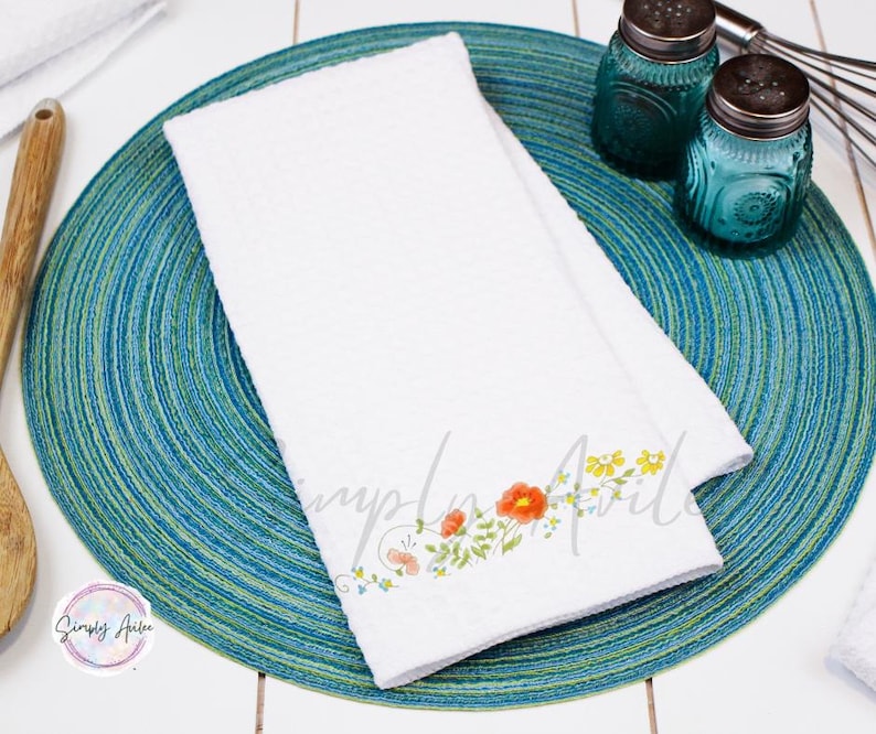 K&ouml;nnte beinhalten: Ein wei&szlig;es K&uuml;chentuch mit einem Blumendesign mit orangefarbenen, gelben und gr&uuml;nen Blumen. Das Tuch liegt auf einem blauen und gr&uuml;nen runden Untersetzer.