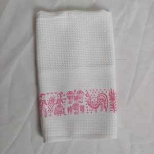 May include: White waffle-weave kitchen towel with a pink border featuring stylized illustrations of people, pineapples, and a rooster. The towel is folded and lies on a white surface.