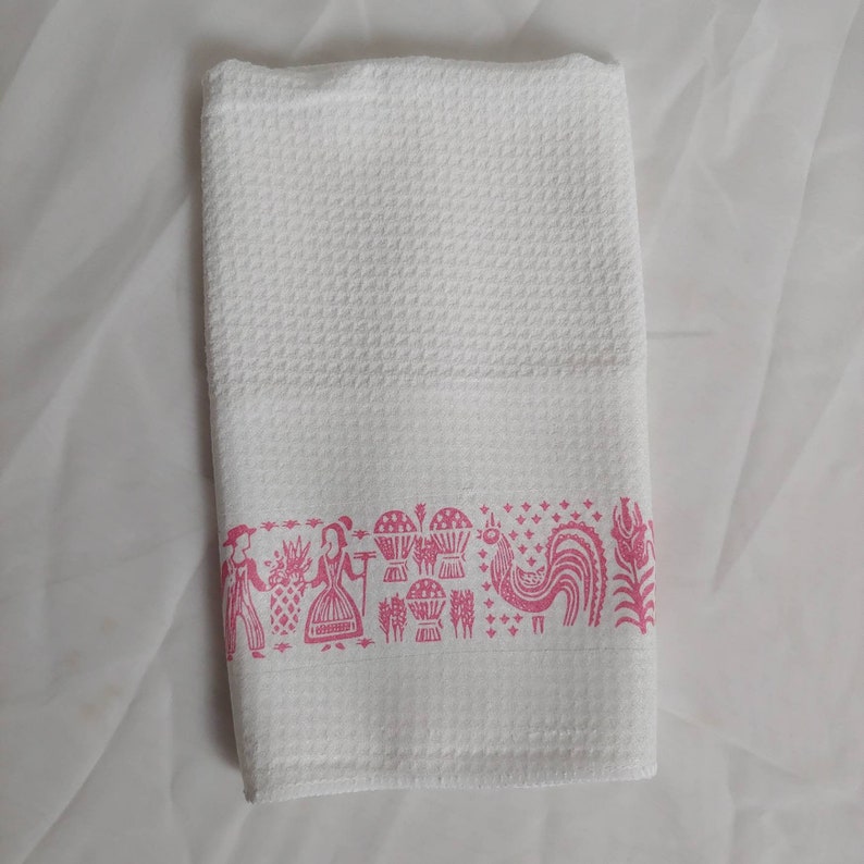 May include: White waffle-weave kitchen towel with a pink border featuring stylized illustrations of people, pineapples, and a rooster. The towel is folded and lies on a white surface.