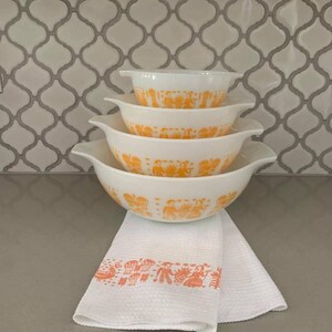 May include: A stack of four white Pyrex bowls with orange designs and handles. A white waffle-weave kitchen towel with an orange border featuring a design of people and objects is beneath the bowls. The background is a white and gray tile pattern.