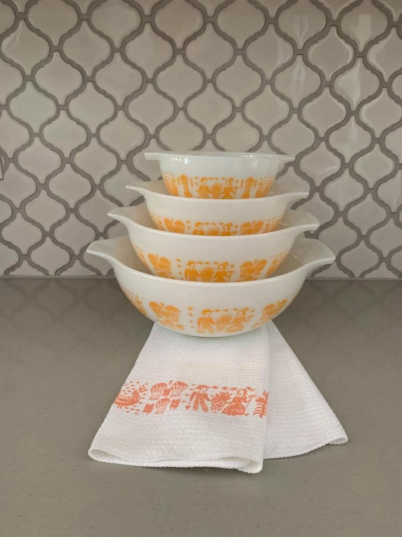 May include: A stack of four white Pyrex bowls with orange designs and handles. A white waffle-weave kitchen towel with an orange border featuring a design of people and objects is beneath the bowls. The background is a white and gray tile pattern.