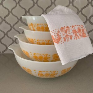 May include: A set of four white mixing bowls with orange decorative patterns. The bowls are stacked, with a white towel featuring an orange print resting on top. The bowls have a classic design, suitable for kitchen use.