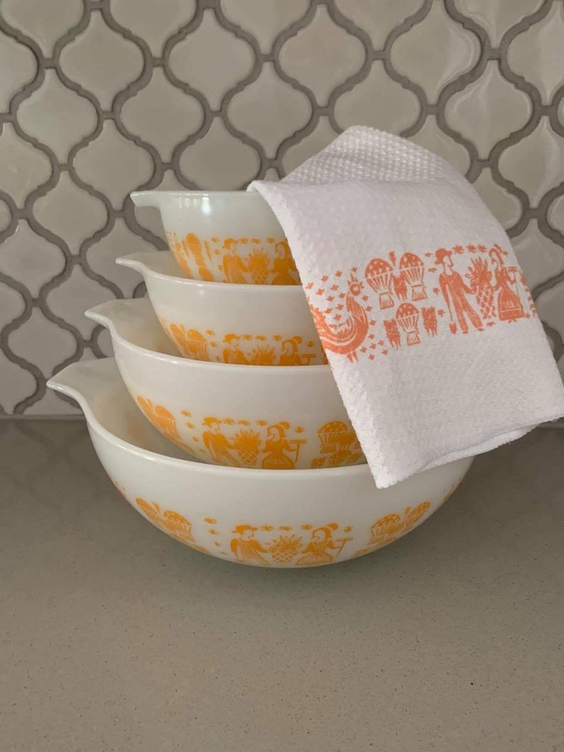 May include: A set of four white mixing bowls with orange decorative patterns. The bowls are stacked, with a white towel featuring an orange print resting on top. The bowls have a classic design, suitable for kitchen use.