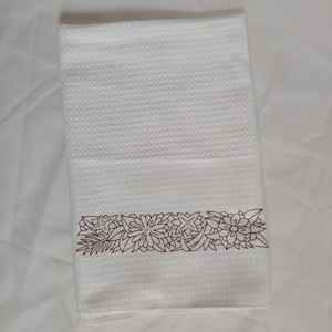 Puede incluir: Toalla de cocina blanca con un borde floral bordado marrón.