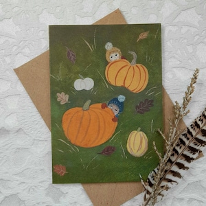 May include: A green card with an autumnal theme featuring two children hiding in pumpkins. The pumpkins are orange and have a green stem. The background is green with brown leaves and white mushrooms.