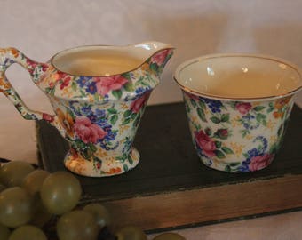 Może przedstawiać: Zestaw vintage z dzbankiem do śmietanki i cukiernicą w kwiatowy wzór. Dzbanek do śmietanki jest biały z kwiatowym wzorem w odcieniach różowego, żółtego, niebieskiego i zielonego. Cukiernica jest biała z podobnym kwiatowym wzorem.