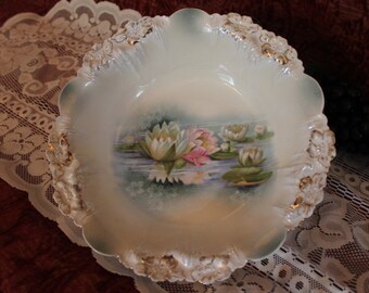 May include: A white porcelain bowl with a scalloped edge and a floral design featuring water lilies in shades of pink, white, and green. The bowl has a gold trim around the edge.