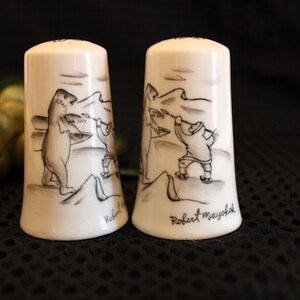 May include: Two white ceramic salt and pepper shakers with black and white illustrations of a bear and a person fishing. The shakers are signed "Robert Mayoh".