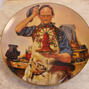 May include: A porcelain plate featuring a painting of a man in a blue shirt and overalls holding a small red Ferris wheel. The plate has a gold rim and the artist's signature "Norman Rockwell" is visible in the bottom right corner.