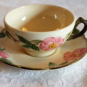 May include: A vintage teacup and saucer set featuring a floral design. The cup and saucer are cream-coloured with pink roses and green leaves.