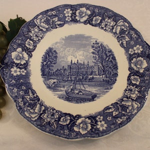 May include: A blue and white ceramic plate with a floral border and a scene of a building with trees and a river in the center.