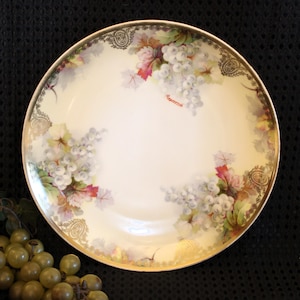 May include: A white porcelain plate with a gold rim and a floral design featuring grapes and leaves. The plate has a cream-coloured background and the grapes are painted in shades of white, pink, and green.