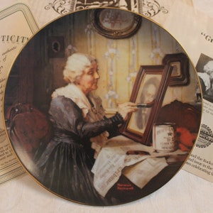 May include: A decorative collector's plate featuring a painting of an older woman painting a portrait of a younger woman in a gold frame. The plate is trimmed in gold and has the words "Grandma's Love" by Norman Rockwell printed on it.