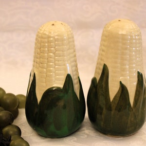 May include: Two ceramic salt and pepper shakers in the shape of corn on the cob. The shakers are cream-coloured with a textured surface, green husks, and small holes at the top. The design is intended to look like a fresh ear of corn.