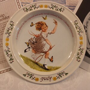 May include: A white ceramic plate with a green vine border and red berries. The center of the plate features a colorful illustration of a girl skipping with a jump rope and butterflies. The text "A certified plate in the original and only Sarah Stilwell Weber Calendar Collection" is printed around the edge of the plate. The word "July" is printed in the center of the plate.