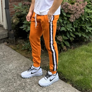 May include: A person wearing a white t-shirt and orange sweatpants with white and brown stripes. The sweatpants have a drawstring waist and the person is wearing white sneakers.