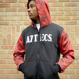 May include: Black zip-up hoodie with red patterned sleeves and hood. The word "AZTECS" is printed in white across the chest. The hoodie has a front zipper and pockets. The person is wearing a white t-shirt underneath.
