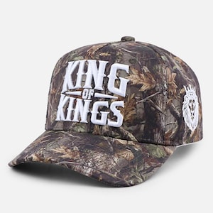 May include: A camouflage baseball cap featuring the white embroidered text KING OF KINGS on the front panel. The side of the cap displays a white embroidered lion head graphic wearing a crown. The fabric has a brown, green, and tan forest pattern.