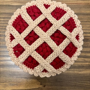 Può includere: Un sottobicchiere fatto a mano a forma di torta all'uncinetto. Il sottobicchiere presenta un ripieno rosso con un motivo a crosta a griglia di colore beige. Il bordo del sottobicchiere è smerlato. Il sottobicchiere è rotondo.