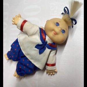 May include: A vintage doll with a round face, blue eyes, and a blonde ponytail tied with a blue bow. The doll wears a white sailor-style shirt with red trim and a blue bow, paired with blue pants with red polka dots.