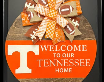 Puede incluir: Un letrero de puerta redondo de madera con acabado en madera oscura y una franja horizontal naranja brillante con el texto blanco "WELCOME TO OUR TENNESSEE HOME" y una letra "T" grande. Un lazo decorativo con patrones de fútbol y cuadros.