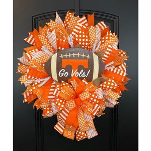 May include: A decorative wreath featuring a football design with the text "Go Vols!". The wreath is made with orange and white ribbons in various patterns, including polka dots and stripes. The wreath is designed for door decor.