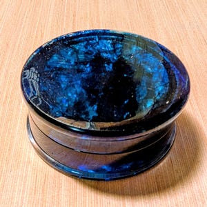 May include: A round, two-piece container with a dark blue, marbled top and a silver-colored base. The top has a glossy finish with swirls of lighter blue and black. The base has a metallic sheen. The container sits on a light brown wooden surface.